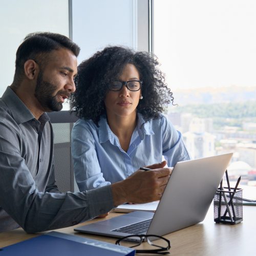 Indian male ceo executive manager mentor giving consultation on financial operations to female African American colleague intern using laptop sitting in modern office near panoramic window.